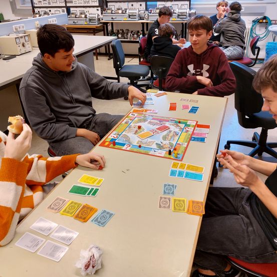 Mehrere Jungen spielen ein Brettspiel in einem Klassenzimmer. Einer von ihnen isst ein Sandwich. Das Spielbrett hat bunte Karten und Würfel.