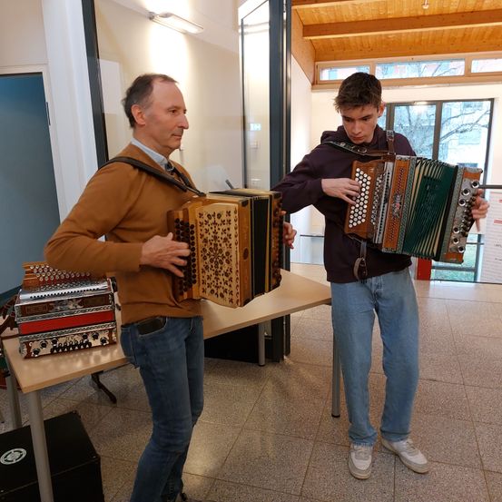 Zwei Männer spielen Akkordeons in einem Raum. Ein Mann ist älter, der andere jung. Sie stehen vor einem Tisch mit anderen Akkordeons darauf.