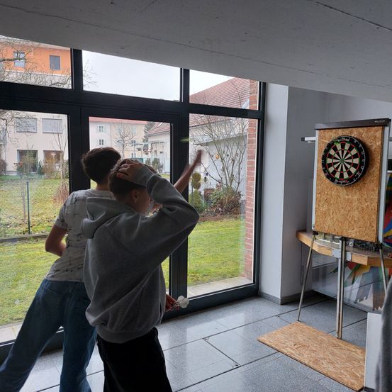 Zwei Jungen spielen Darts drinnen. Einer zielt, während der andere von hinten zuschaut. Ein Dartboard ist an der Wand montiert, und ein Tisch mit einem farbenfrohen Gemälde ist in der Nähe.