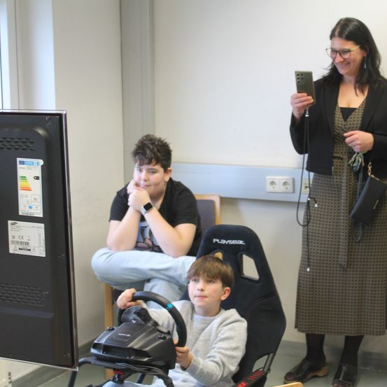 Zwei Kinder spielen an einem Rennsimulator. Ein Junge sitzt in einem Rennsitz, während ein anderer Junge auf seinem Schoß sitzt. Eine Frau hält ein Smartphone vor ihnen.