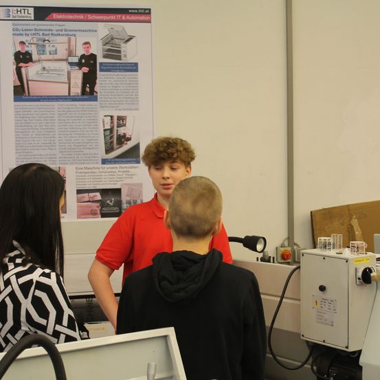 Drei Personen befinden sich in einem Raum mit technischer Ausrüstung. Ein Mann in einem roten T-Shirt spricht mit zwei anderen. Dahinter ist ein Poster an der Wand befestigt.