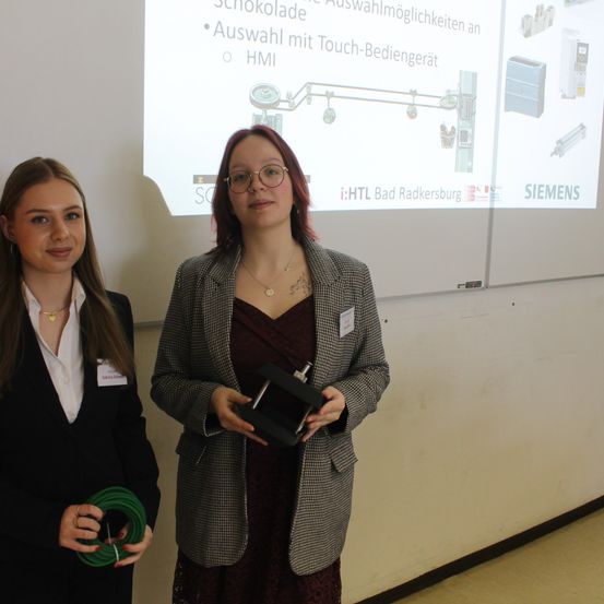 Zwei Frauen stehen vor einer Tafel. Eine hält ein grünes Kabel, die andere ein schwarzes Gerät. Sie tragen beide eine Brille. Der Hintergrund ist eine Projektionsfläche mit einem Diagramm.