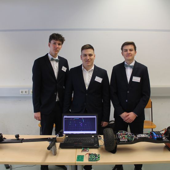 Drei junge Männer in Anzügen und Fliegen stehen hinter einem Tisch mit Laptop und Elektro-Skateboard. Sie scheinen ein Projekt zu präsentieren.