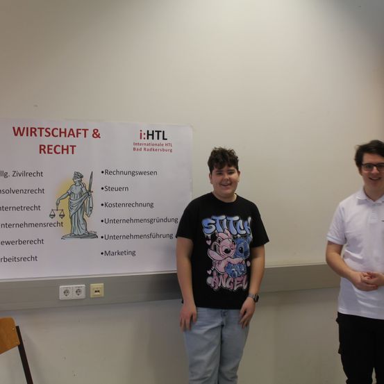 Zwei junge Männer stehen vor einer Wand mit einem Plakat mit der Aufschrift 'Wirtschaft & Recht'. Der Mann auf der linken Seite trägt ein schwarzes T-Shirt und Jeans, der Mann auf der rechten Seite trägt ein weißes Polohemd und eine Brille.
