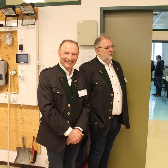 Zwei Männer in Uniformen stehen nebeneinander in einem Raum mit Holzwänden und elektrischen Geräten. Einer lächelt und der andere hat einen Namensschild.