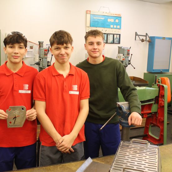 Drei junge Männer in einer Werkstatt, zwei in roten Hemden und einer in grün, stehen neben einer Maschine. Einer hält ein Metallobjekt, wahrscheinlich eine Uhr. Hinter ihnen befinden sich Werkzeuge und Ausrüstung.