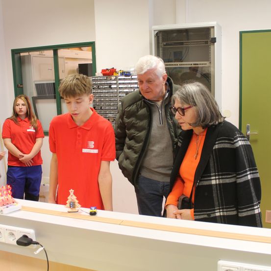 Ein Junge in einem roten Shirt untersucht eine kleine Modelleisenbahn auf einer Strecke, während Erwachsene zuschauen.