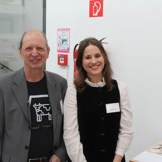 Ein Mann und eine Frau stehen lächelnd vor einer weißen Wand mit Feuerlöscher und Sicherheitsschild.