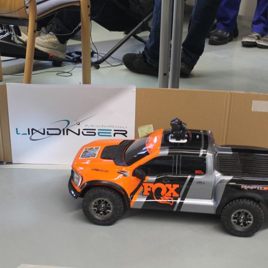 Ein orange-grauer Spielzeug-LKW mit FOX-Aufklebern steht auf einem grauen Boden. Dahinter befindet sich ein Karton mit einem weißen Schild, auf dem LININGER Modellbau steht.