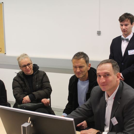 Fünf Personen in einem Raum, drei von ihnen sitzen auf Stühlen, während zwei stehen. Der Mann vor ihnen lächelt in die Kamera. Ein Computermonitor steht vor ihnen.