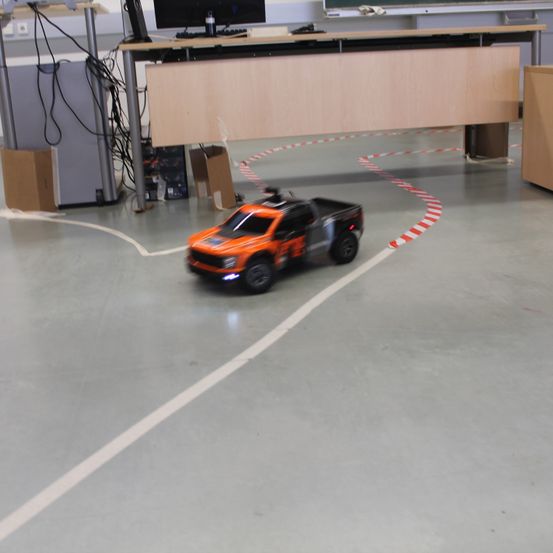 Ein ferngesteuerter orangefarbener und schwarzer LKW fährt auf einer mit weißen und roten Linien markierten Spur. Der LKW befindet sich in einem Raum mit einem Holzschreibtisch und einem schwarzen Computermonitor.