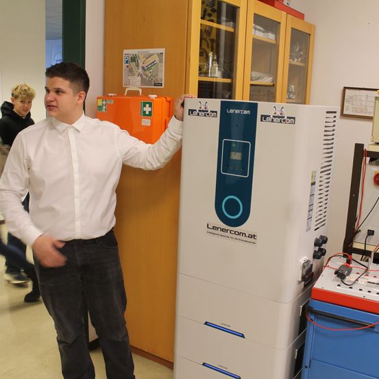 Ein Mann in einem weißen Hemd steht neben einer weißen Maschine mit der Aufschrift Lehercom in einem Raum mit einem hölzernen Schrank und einer orangefarbenen Erste-Hilfe-Box.