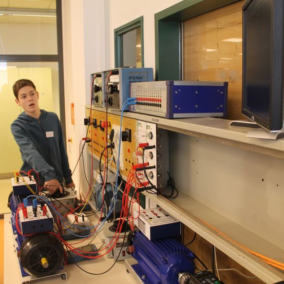 Ein junger Mann stellt elektrische Geräte in einer Werkstatt ein. Er steht vor einem Regal, das verschiedene Geräte enthält, darunter eine blau-silberne Maschine mit mehreren Drähten und Tasten.
