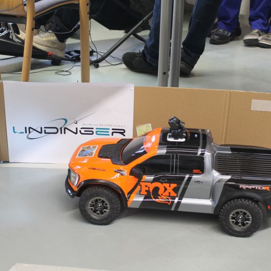 Ein orange-schwarzes Spielzeugauto mit dem Wort 'FOX' an der Seite steht in einem Raum. Ein Schild mit dem Wort 'LINDINGER' ist im Hintergrund zu sehen. Einige Personen stehen hinter dem Auto.