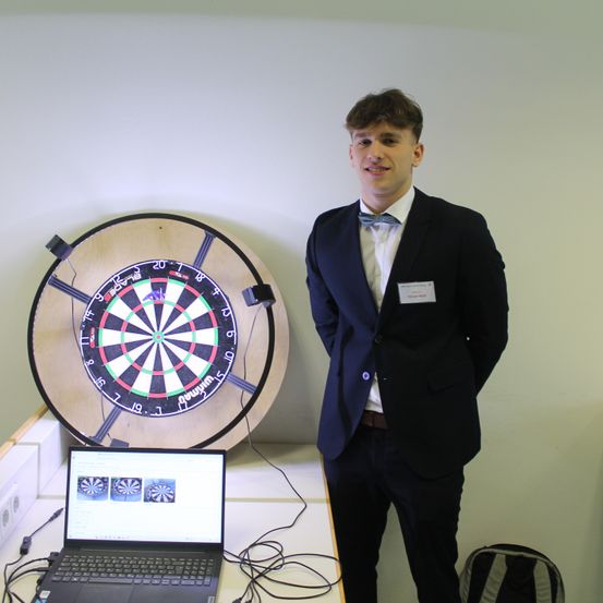 Ein Mann in Anzug steht neben einem großen elektronischen Dartboard und einem Laptop auf einem Schreibtisch.