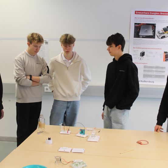 Drei junge Männer stehen um einen Tisch mit wissenschaftlicher Ausrüstung. Sie befinden sich in einem Raum mit einer weißen Wand, einem Plakat und einer Tafel. Auf dem Tisch stehen verschiedene Gegenstände, darunter eine Glasflasche.