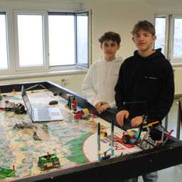 Zwei Jungen stehen neben einem großen Tisch voller Legos und einem Laptop. Einer trägt eine weiße Kapuzenjacke, der andere eine schwarze. Hinter ihnen befinden sich Fenster und ein weißer Heizkörper.