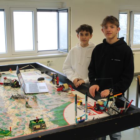Zwei Jungen stehen neben einem großen Tisch voller Legos und einem Laptop. Einer trägt eine weiße Kapuzenjacke, der andere eine schwarze. Hinter ihnen befinden sich Fenster und ein weißer Heizkörper.