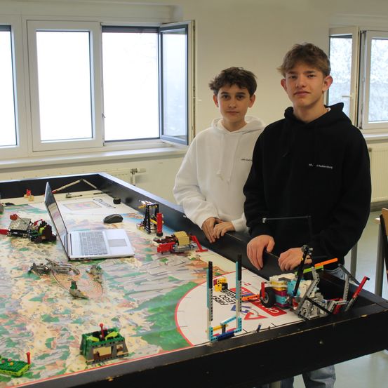 Zwei Jungen stehen neben einem großen Tisch voller Legos und einem Laptop. Einer trägt eine weiße Kapuzenjacke, der andere eine schwarze. Hinter ihnen befinden sich Fenster und ein weißer Heizkörper.