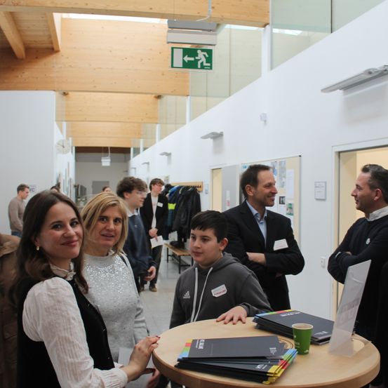 Eine Gruppe von Erwachsenen und Kindern versammelt sich um einen Tisch mit Ordnern und Büchern. Der Raum hat Holzbalken, weiße Wände und eine Glasdecke. Ein Ausfahrtsschild ist über ihnen.