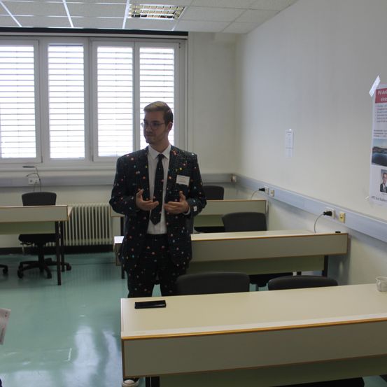 Ein Mann in einem Anzug steht in einem Klassenzimmer und gestikuliert mit den Händen. Hinter ihm sind Reihen von Tischen und Stühlen zu sehen, mit einer Tafel an der Wand.