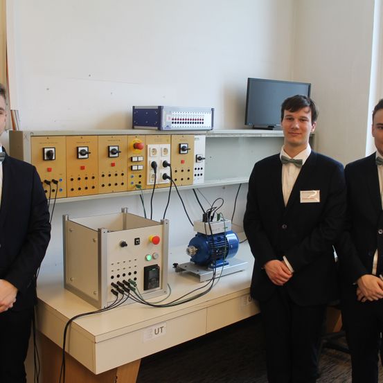 Drei Männer in Anzügen stehen vor einem weißen Tisch mit einem mechanischen Gerät, einem Monitor und einem Schaltpult auf einem Regal.