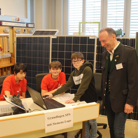 Drei Jungen sitzen an einem Schreibtisch mit einem Laptop, während ein Mann in Uniform hinter ihnen steht und lächelt. Dahinter sind Solarmodule an der Wand montiert.