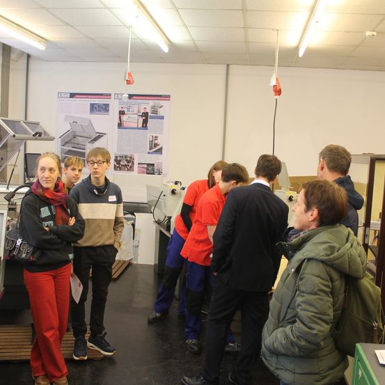 In einer Werkstatt stehen eine Gruppe von Schülern und Erwachsenen um eine Maschine herum, einige schauen auf ein Poster an der Wand.