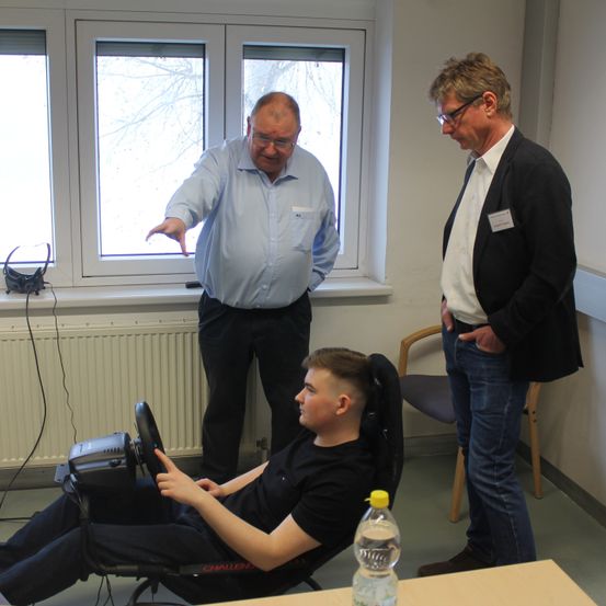 Drei Männer in einem Raum mit Fenstern. Ein Mann in einem blauen Hemd gestikuliert während er spricht. Ein anderer Mann in einem schwarzen Anzug steht mit den Händen in den Taschen. Ein dritter Mann in einem schwarzen T-Shirt sitzt in einem Stuhl mit einem Lenkrad.