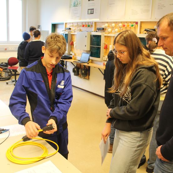 In einem Workshop arbeitet ein junger Mann in einer blauen Jacke an einem gelben Kabel. In der Nähe beobachtet ihn ein Mädchen mit Brille und einer schwarzen Kapuzenjacke. Andere Leute sind im Hintergrund.