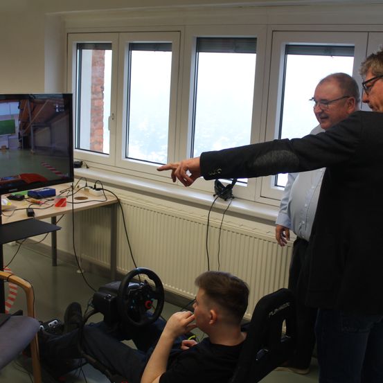 Drei Männer sind in einem Raum mit einem Monitor, einem Lenkrad und einem Heizkörper. Ein Mann sitzt auf einem Stuhl, während ein anderer auf den Monitor zeigt.