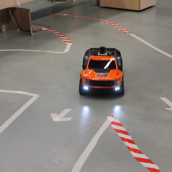 Ein orangefarbenes Spielzeugauto fährt auf einem Boden mit weißen und roten Streifen, beleuchtet von Scheinwerfern.