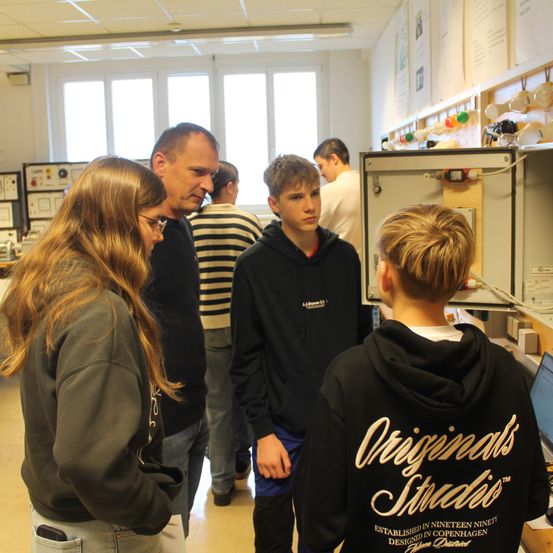 Eine Gruppe von Menschen steht in einem Raum mit einem Monitor auf dem Schreibtisch, und eine Person mit Brille steht in der Nähe des Monitors.