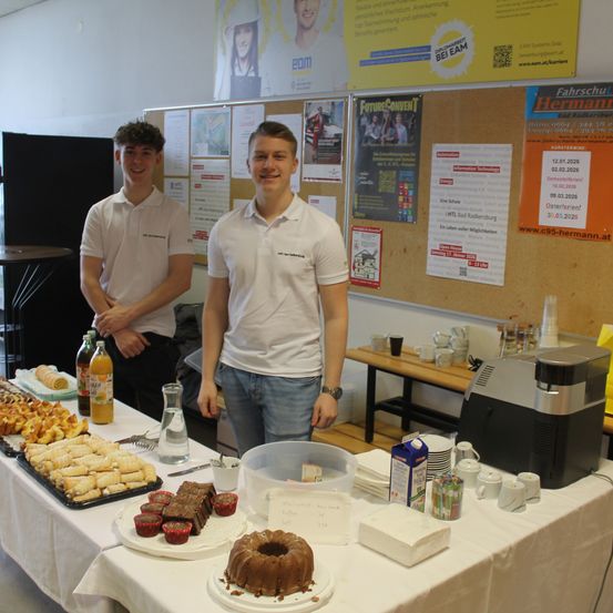 Zwei junge Männer stehen hinter einem Tisch mit einer Vielzahl von Desserts, Getränken und Tassen. Ein Mann lächelt, während der andere nach unten schaut. Hinter ihnen befindet sich eine Wand mit verschiedenen Postern und Werbung.