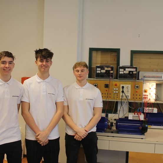 Drei junge Männer in weißen Poloshirts stehen in einem Labor, lächeln und posieren für ein Foto. Hinter ihnen befindet sich eine weiße Wand mit verschiedenen elektrischen Geräten.