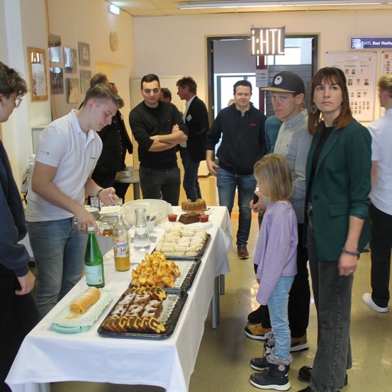 Eine Gruppe von Menschen versammelt sich um einen Tisch mit Essen, möglicherweise auf einer Party. Eine Frau mit Armband steht neben einem kleinen Mädchen.