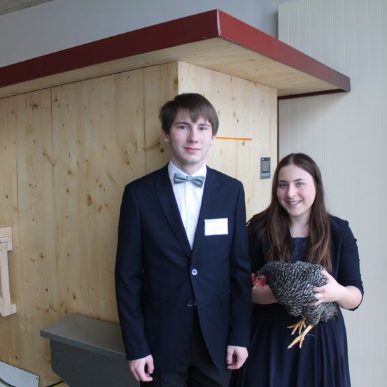 Ein junger Mann und eine junge Frau stehen nebeneinander und lächeln, mit einer Holzwand im Hintergrund. Der Mann trägt eine Fliege und die Frau hält ein Huhn.