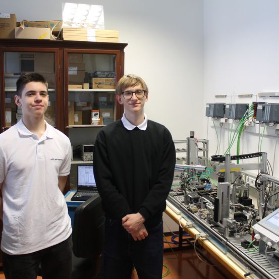 Zwei junge Männer stehen in einem mit wissenschaftlichen Geräten gefüllten Raum. Einer trägt ein weißes Hemd, der andere ein schwarzes. Hinter ihnen steht ein Holzschrank und ein Computer.