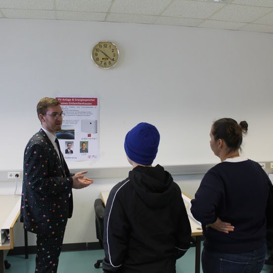 Ein Mann in einem Anzug spricht mit zwei Personen in einem Klassenzimmer. An der Wand hängt ein Plakat mit Text auf Deutsch und einer Uhr darüber.
