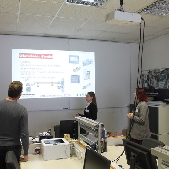 Drei Personen stehen in einem Raum vor einer Projektionsleinwand, auf der der Text 'Schokoladen Gondel' zu sehen ist. Zwei Frauen hören der Präsentation zu. Ein Mann schaut auf den Bildschirm.