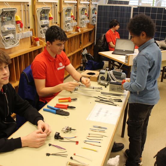 Mehrere Schüler in einem Klassenzimmer, einer hält ein Werkzeug und ein anderer beobachtet. Sie sind von verschiedenen Werkzeugen und Kabeln auf einem Tisch umgeben.