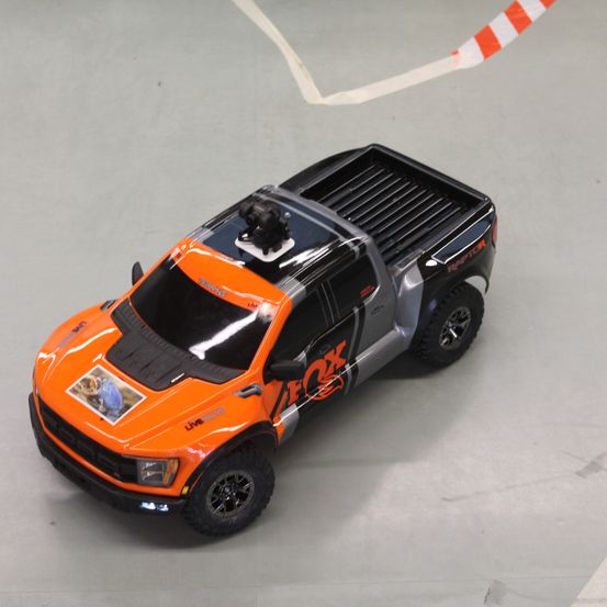 Ein orangefarbener und schwarzer Truck mit 'FOX' auf der Seite steht auf einem Betonboden. Der Truck hat ein großes Bild auf der Vorderseite.