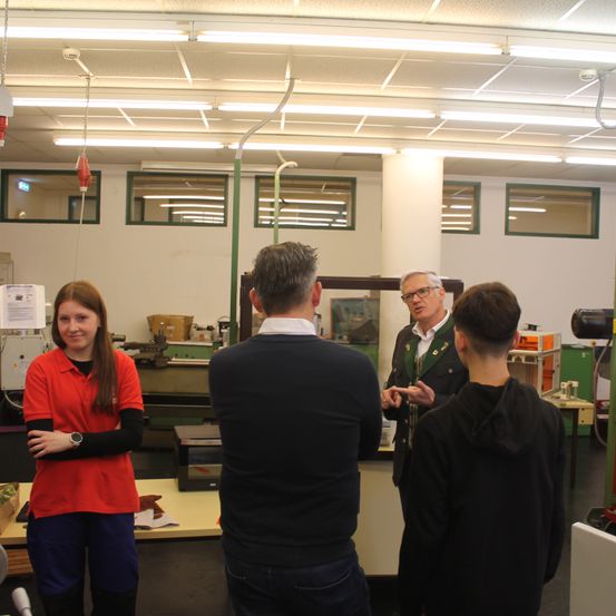 Eine Gruppe von Menschen befindet sich in einer Werkstatt. Ein Mann mit Brille spricht mit zwei anderen. Eine Frau in einem roten Shirt beobachtet. Ausrüstung und Fenster sind im Hintergrund.