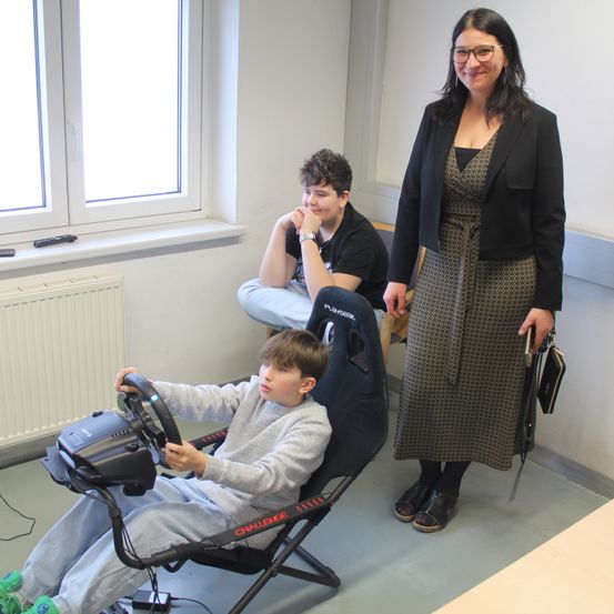 Drei Personen in einem Raum mit einem Fenster. Einer sitzt in einem Gaming-Stuhl, ein anderer sitzt auf einem Stuhl und eine Frau steht vor ihnen.