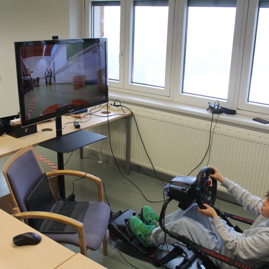 Ein junger Mensch sitzt in einem Rennsitz vor einem Monitor in einem Klassenzimmer. Auf dem Stuhl befindet sich ein Laptop und auf dem Schreibtisch eine Maus. Der Monitor zeigt eine virtuelle Umgebung.