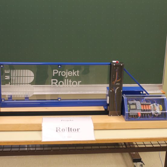 Ein Projekt namens Rolltor, ausgestellt auf einem Holztisch, mit einer blauen Struktur und elektronischen Komponenten. Der Hintergrund ist eine grüne Tafel mit weißen Punkten.