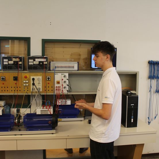 Ein junger Mann steht in einem Labor vor einem Schreibtisch mit einem Computer und verschiedenen Maschinen darauf.