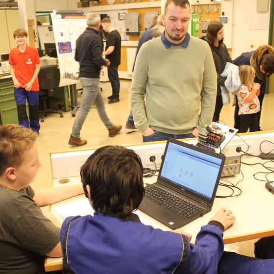 Zwei Jungen arbeiten an einem Laptop, während andere zusehen, ein Mann lächelt im Hintergrund.