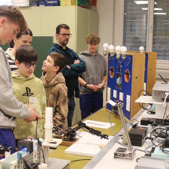Eine Gruppe von Schülern betrachtet eine Maschine mit Lichtern darauf in einem Klassenzimmer.
