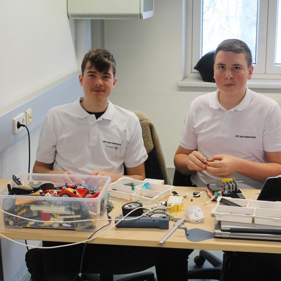 Zwei junge Männer sitzen an einem Schreibtisch mit verschiedenen Elektronikgeräten. Einer hält ein Werkzeug, der andere einen Laptop. Sie tragen beide weiße Poloshirts.